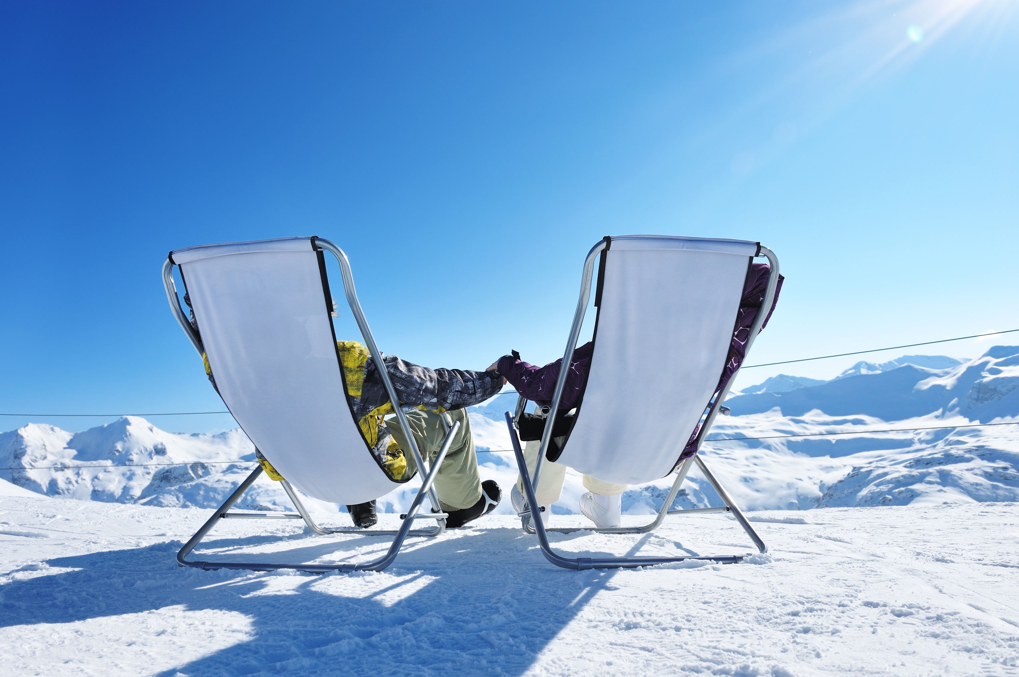 Two people in lounge chairs on a snowy mountain peak, holding hands and looking at the view
